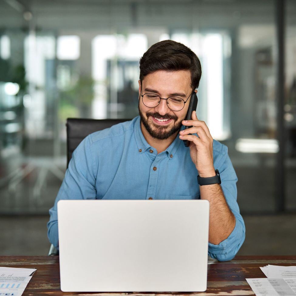 Smiling businessman making a call on mobile phone while looking at his laptop.