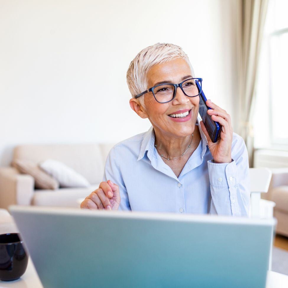 Smiling advisor with white hair working on laptop while talking on a mobile phone.