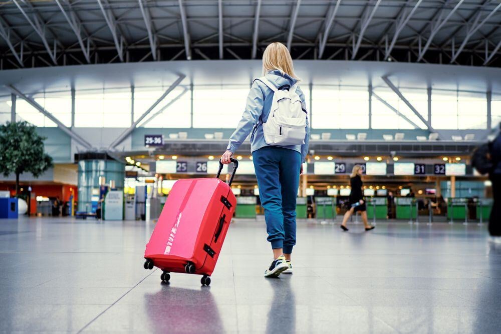 Young woman in casual wear standing in an international airport terminal with a pink suitcase.